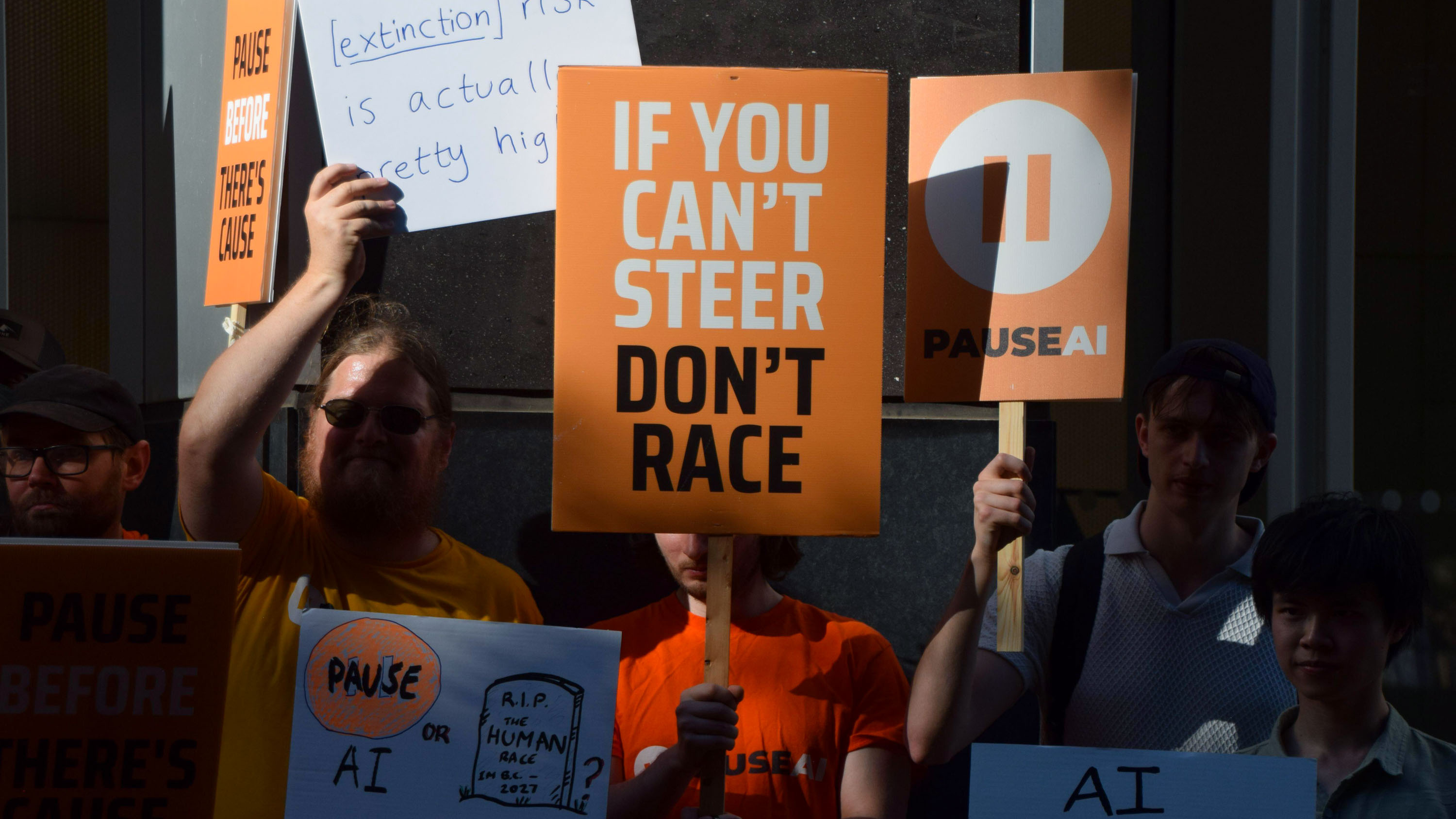 Close-up of a crowd of protestors holding orange Pause AI signs including one that reads "If you can't steer don't race" as well as others partially visible which mention extinction and a gravestone with RIP human race.