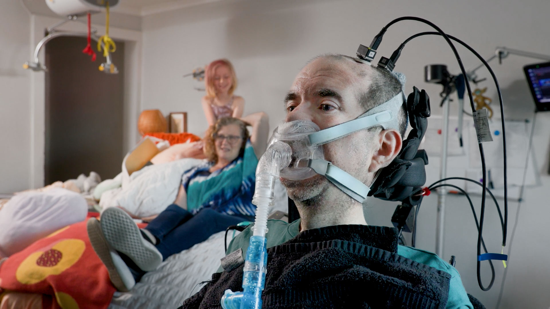 Casey Harrell in the foreground with the BCI implants and cables protruding from his head. His wife and young daughter are sitting in the background